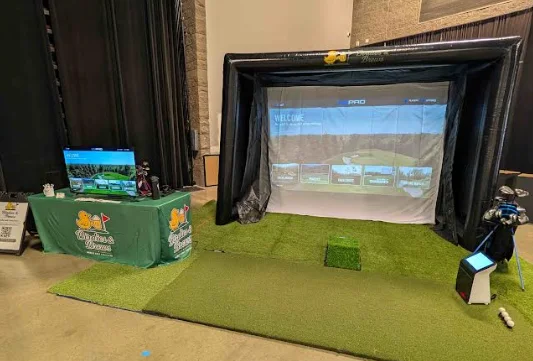The compact Birdies & Brews simulator bay set up inside an event hall beside a branded table and screen.
